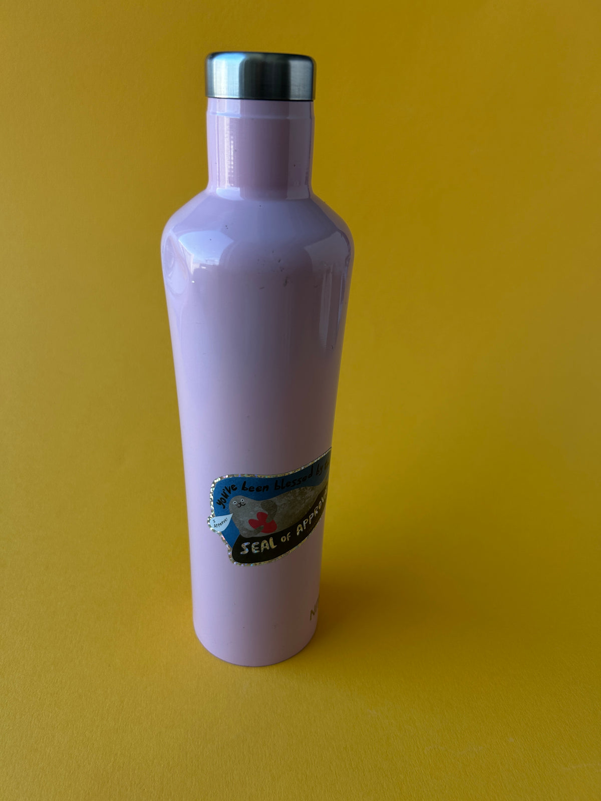 A light purple plastic bottle with a silver metal cap, lying horizontally on a solid yellow background. A blue and white label is visible on the side of the bottle.