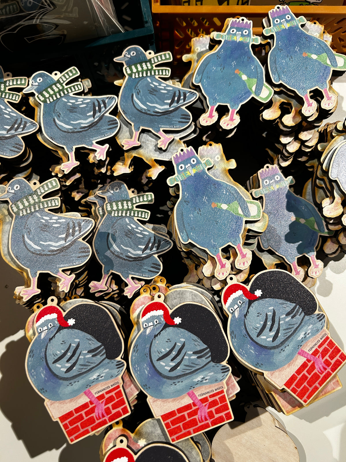 A close-up of multiple decorative pigeon-shaped ornaments or charms displayed together. The designs include blue pigeons with pink feet, some wearing green-and-white striped scarves, and several with red Santa hats above red brick fireplace shapes.