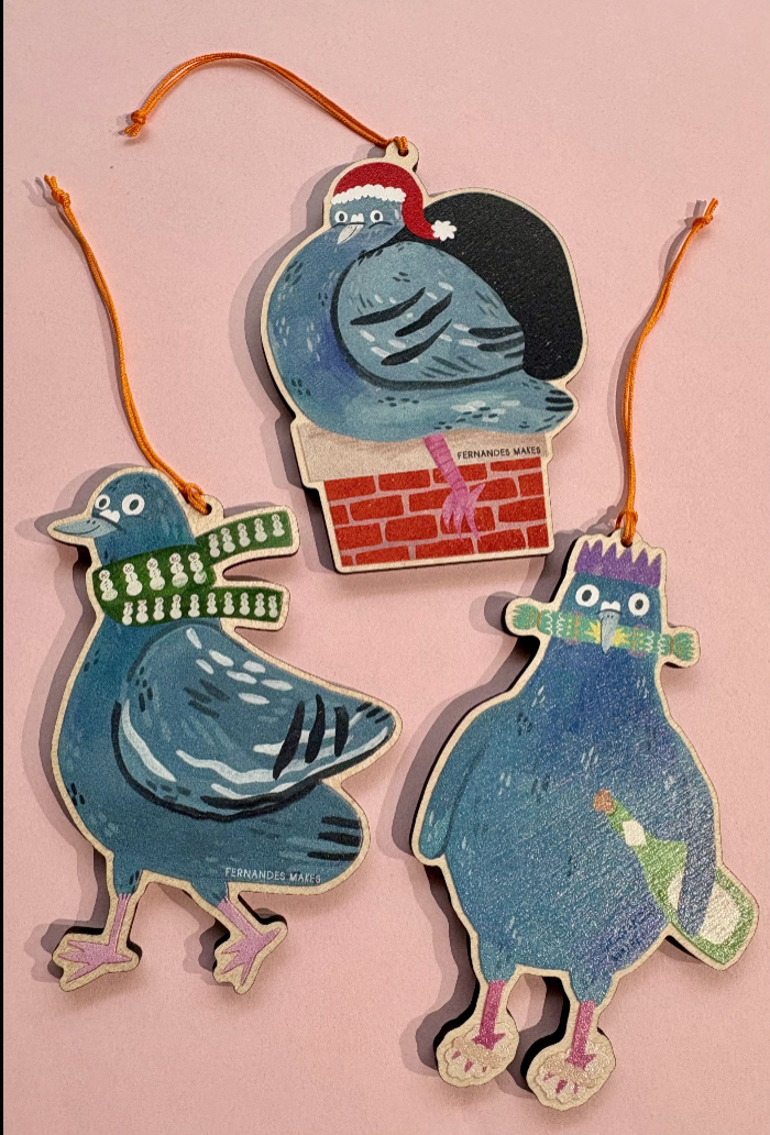 Three whimsical hand-painted wooden Christmas ornaments shaped like pigeons, displayed on a soft pink background. The ornaments feature colorful winter-themed illustrations, including one with a green scarf, one with a red Santa hat and brick chimney, and one with purple hair and a green scarf.