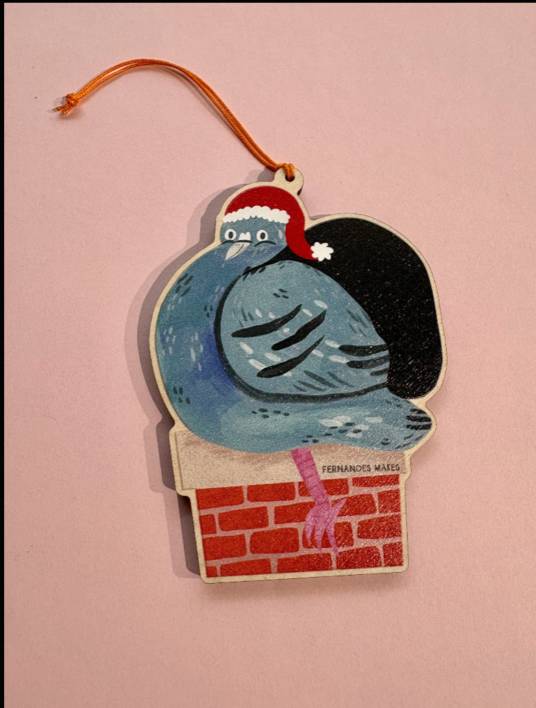 A hand-painted wooden Christmas ornament featuring a blue pigeon wearing a red Santa hat and perched on a red brick chimney. The ornament has a gold hanging loop at the top and is displayed against a solid pink background.