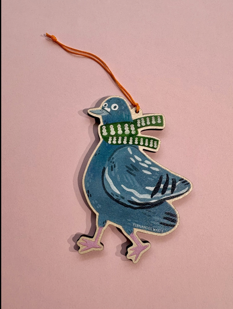 A hand-painted wooden Christmas ornament shaped like a blue pigeon wearing a green and white striped scarf, hanging from an orange string against a solid pink background.