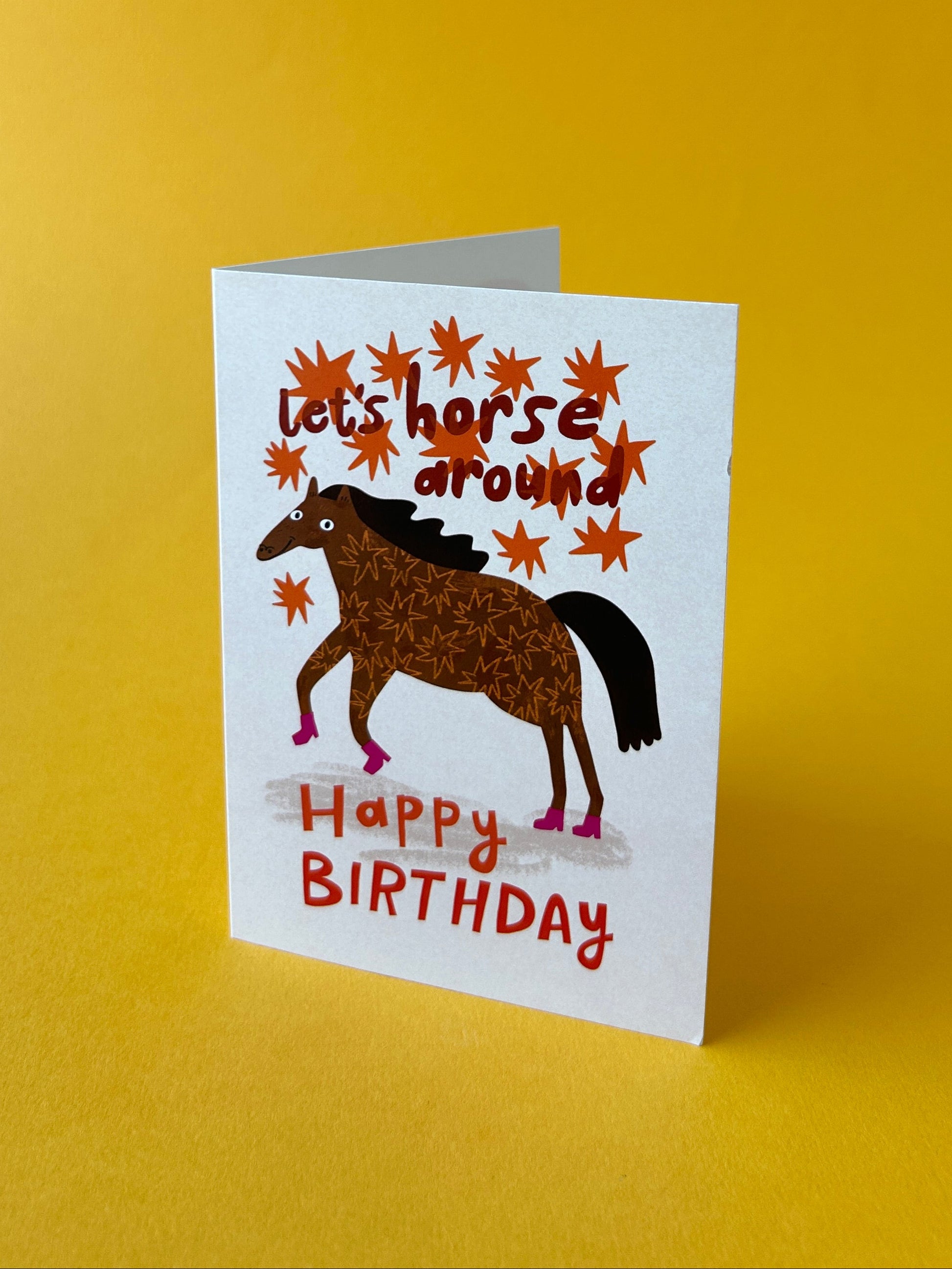 A white square birthday card with a cartoon horse illustration and the text 'Let's horse around Happy Birthday', standing on a yellow background.