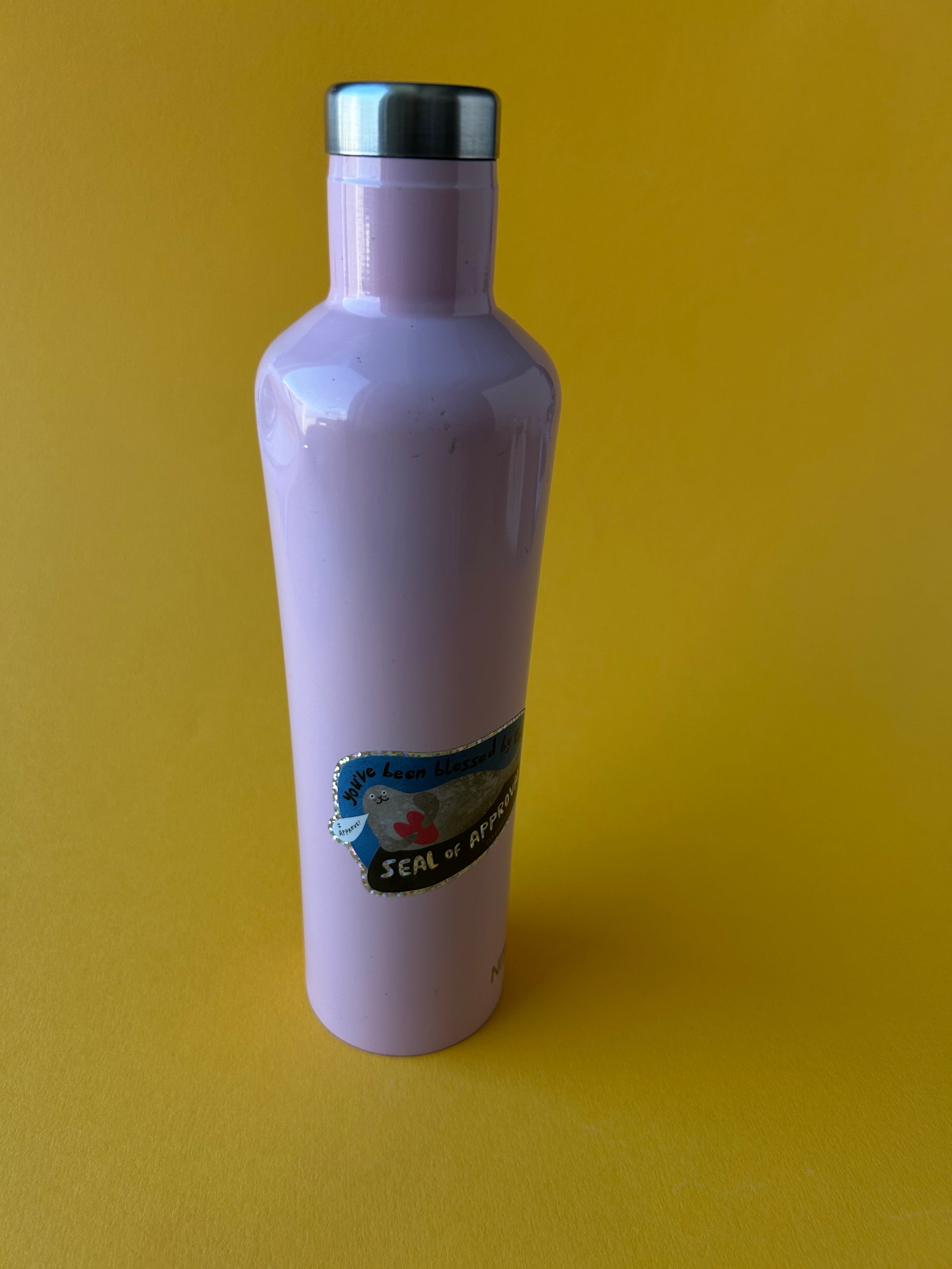 A light purple plastic bottle with a silver metal cap, lying horizontally on a solid yellow background. A blue and white label is visible on the side of the bottle.