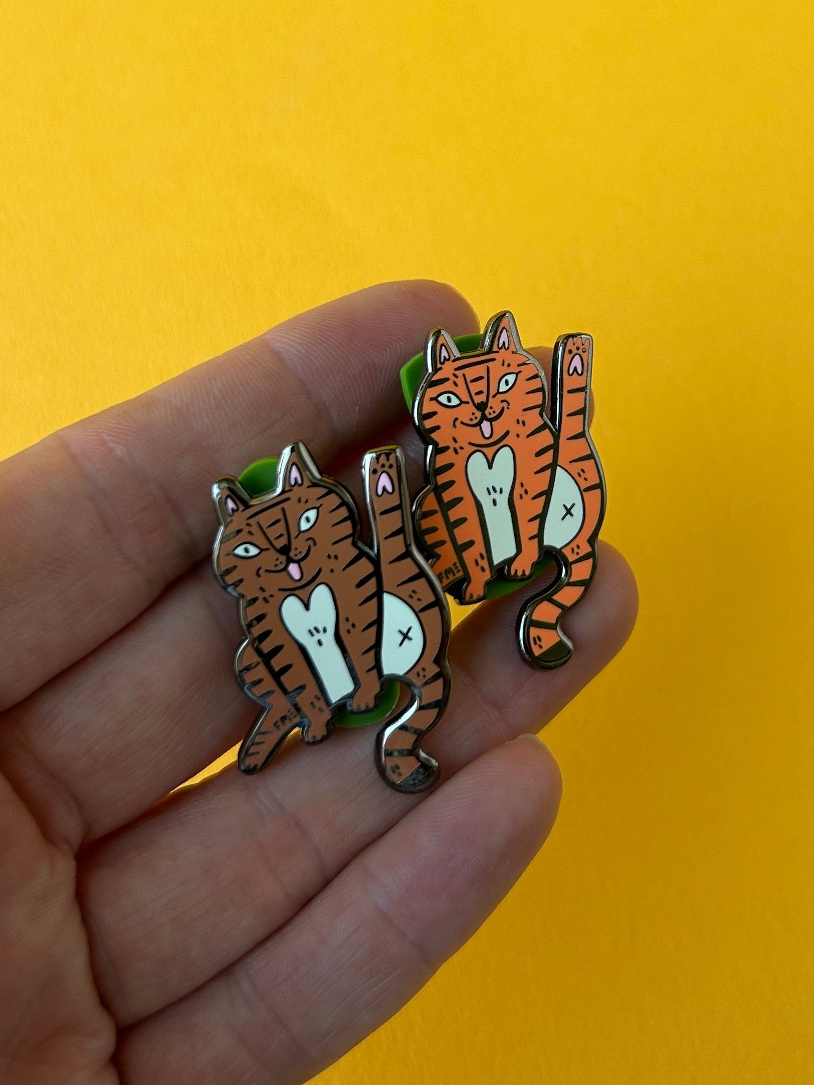 A hand holding two small tiger-shaped enamel pins against a solid yellow background.