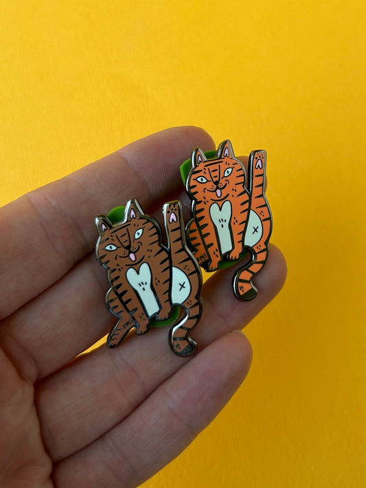 A hand holding two small tiger-shaped enamel pins against a solid yellow background.