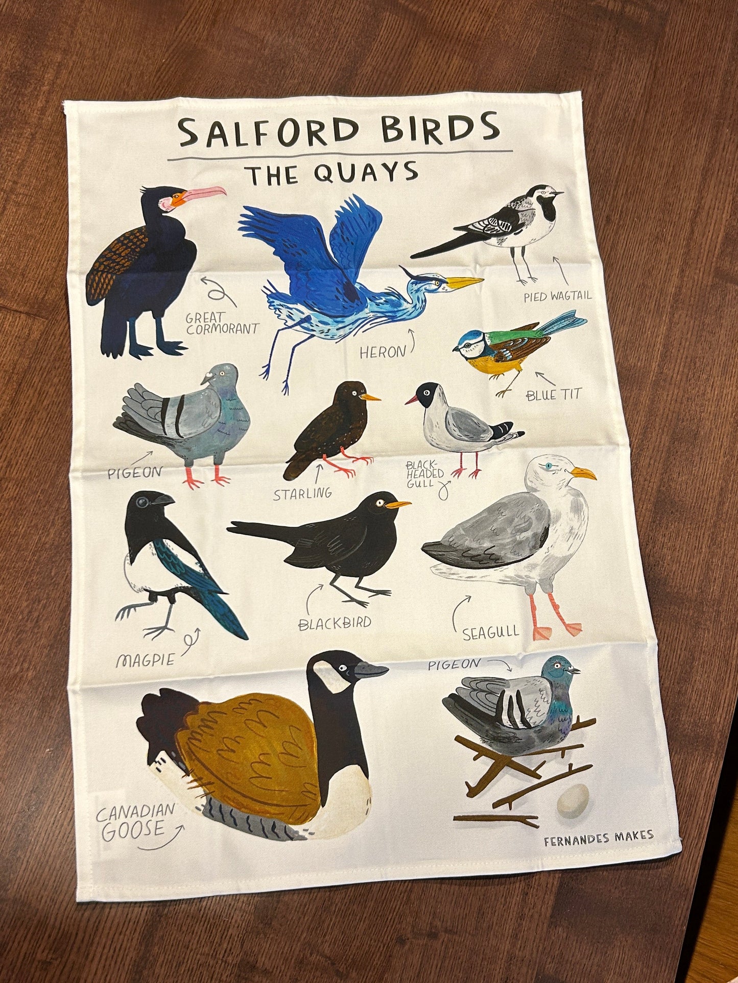 A printed tea towel featuring illustrated birds of 'Salford Birds The Quays' by Fernandes Makes, laid flat on a dark wood table.