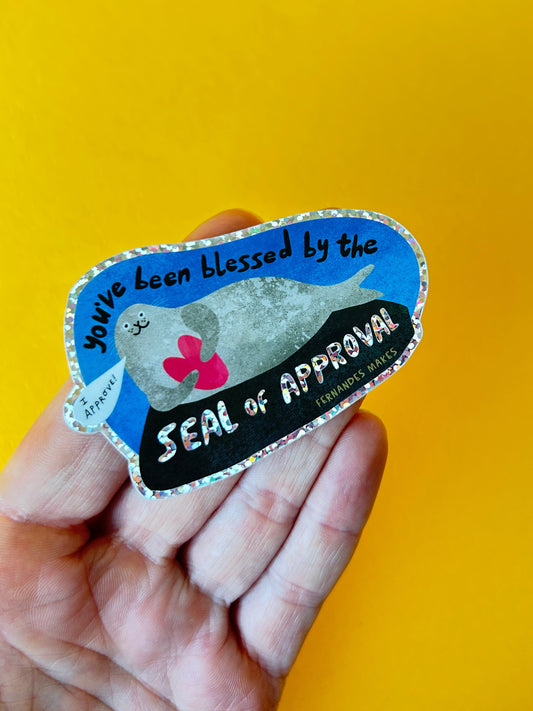 A hand holding a die-cut sticker with a blue, black, white, and pink design featuring the text 'You've been blessed by the Seal of Approval' on a solid yellow background.