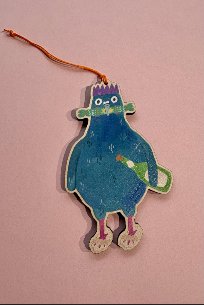 A hand-painted wooden hanging ornament shaped like a pigeon with a purple crown, green cracker and holding a green bottle, suspended by an orange string on a pink background.